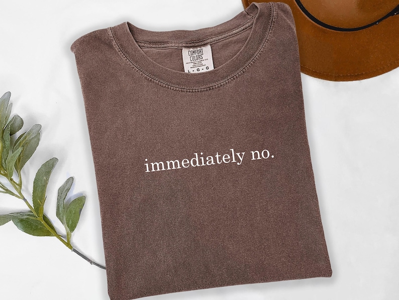 Op de afbeelding: Een bruin Comfort Colors T-shirt met de witte tekst "immediately no.". Het shirt ligt opgevouwen op een witte ondergrond, met een bruine hoed op de achtergrond. Een takje groene bladeren bevindt zich in de linker benedenhoek.