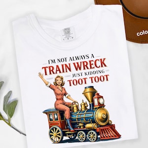 May include: White t-shirt with a retro illustration of a woman on a train. The text reads "I'm not always a train wreck just kidding toot toot." The design features a red, blue, and gold train with a woman in an orange jumpsuit.