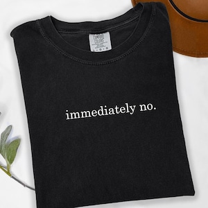 Op de afbeelding: Zwart T-shirt met de witte tekst "immediately no" in het midden. Het shirt is opgevouwen en ligt op een wit oppervlak, met een bruine hoed op de achtergrond. Een takje groen is te zien in de linker benedenhoek.