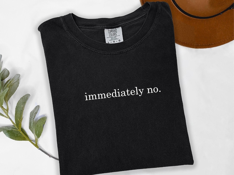 Op de afbeelding: Zwart T-shirt met de witte tekst "immediately no" in het midden. Het shirt is opgevouwen en ligt op een wit oppervlak, met een bruine hoed op de achtergrond. Een takje groen is te zien in de linker benedenhoek.