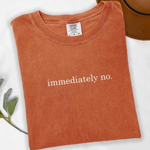 Op de afbeelding: Oranje-bruin T-shirt met de witte tekst "immediately no.". Het shirt is van het merk Comfort Colors, met een ronde hals en korte mouwen. Een bruine hoed is gedeeltelijk zichtbaar op de achtergrond.