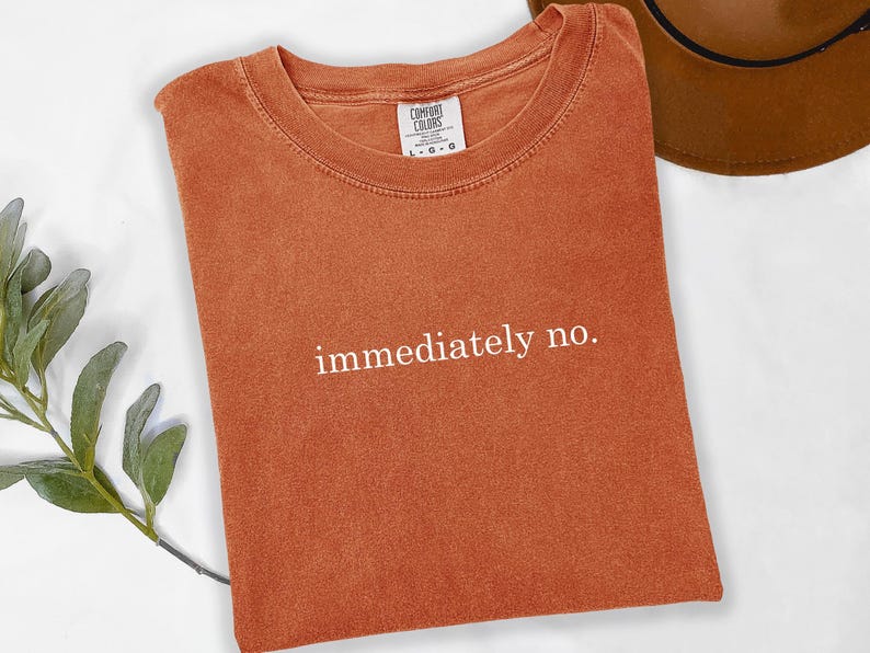 Op de afbeelding: Oranje-bruin T-shirt met de witte tekst "immediately no.". Het shirt is van het merk Comfort Colors, met een ronde hals en korte mouwen. Een bruine hoed is gedeeltelijk zichtbaar op de achtergrond.