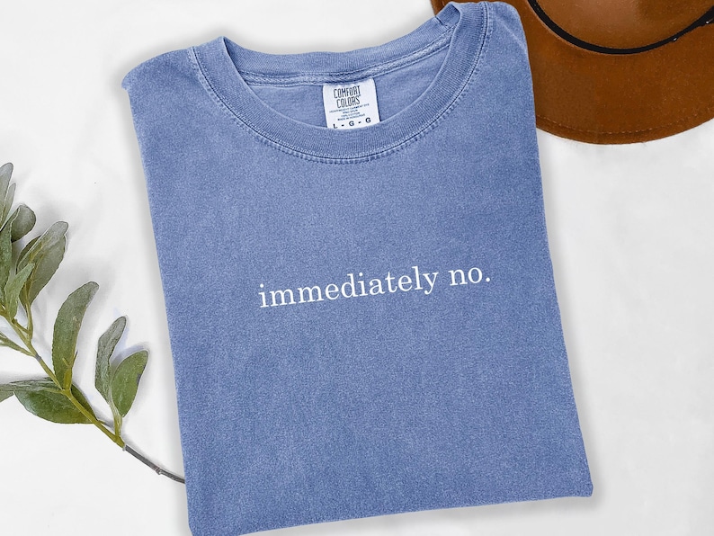 Op de afbeelding: Blauw t-shirt met de woorden "immediately no" in witte letters. Het shirt is opgevouwen en heeft een label met "Comfort Colors". Een bruine hoed is gedeeltelijk zichtbaar op de achtergrond.