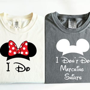 Peut inclure: Deux t-shirts pliés, un crème et un gris. Le t-shirt crème présente un design de Minnie Mouse avec un nœud à pois rouges et les mots "I Do". Le t-shirt gris dit "I Don't Do Matching Shirts."