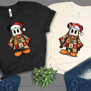 May include: Two t-shirts, one black and one cream, each featuring a cartoon character dressed as a ghost in a Christmas-themed patchwork quilt. The character wears a Santa hat and orange shoes. The shirts are paired with blue jeans.