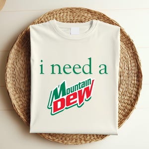 May include: Cream-colored t-shirt with the text "i need a Mountain Dew" in green and red. The Mountain Dew logo is prominently displayed. The shirt is laid on a woven basket.