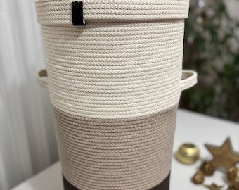 May include: A tall cylindrical storage basket made of coiled rope featuring a three-tone color scheme of cream, light brown, and dark brown. The basket includes a matching lid with a small loop handle, two side handles, and a black leather tab.