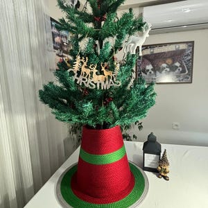 Könnte beinhalten: Ein kleiner künstlicher grüner Weihnachtsbaum, geschmückt mit einem Holzornament mit Hirschen und dem Schriftzug CHRISTMAS. Der Baumfuß ist mit einer rot-grünen gehäkelten Weihnachtsbaumdecke mit grauem Rand bedeckt, die auf einer weißen Fläche steht.