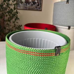 May include: A bright green woven rope basket with a white plastic inner liner. The basket features a horizontal red stitched accent line near the top rim and a small black leather-like tab with a metallic rivet attached to the side.