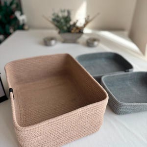 Pode incluir: Três cestos de armazenamento retangulares feitos de corda enrolada. Um cesto grande é bege claro, enquanto dois cestos menores são cinza escuro. Cada cesto apresenta um design simples de topo aberto com uma pequena aba de tecido preto na lateral.