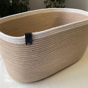 Puede incluir: Una cesta de almacenamiento ovalada construida con cuerda de color canela enrollada y un borde de cuerda blanca. Una pequeña lengüeta rectangular de aspecto de cuero negro está unida al borde superior con un remache metálico y tiene texto.