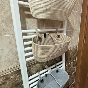 May include: Three woven rope baskets in beige and gray hang from a white towel rack. The baskets are oval-shaped with handles and are designed for bathroom storage. The rack is mounted on a tiled wall.