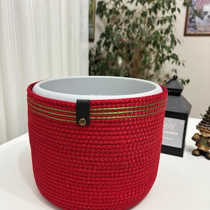May include: A cylindrical planter featuring a vibrant red woven rope exterior with two horizontal lime green stitched lines near the top rim. A small black leather-like tab with a brass-colored metal rivet is attached to the upper edge.