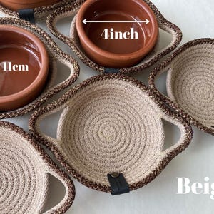 Puede incluir: Un conjunto de cuencos redondos de terracota en soportes de cuerda trenzada beige y marrón con detalles de cuero negro. Los cuencos miden 11 cm de alto y 4 pulgadas de diámetro. Los soportes de cuerda tienen asas integradas y el texto Beige.