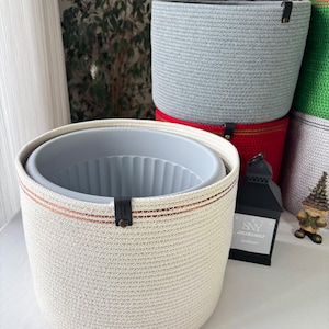 May include: A cylindrical off-white woven rope basket with a thin orange horizontal stripe and a black leather tab featuring a metal rivet. Inside sits a light gray plastic plant pot with vertical ribbed interior walls.