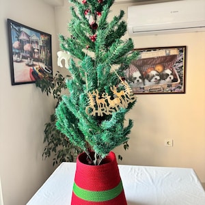 May include: A green artificial Christmas tree stands in a red and green crocheted basket. The tree is decorated with red ornaments and a wooden cutout sign featuring reindeer silhouettes and the word CHRISTMAS in capital letters.