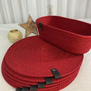 May include: A collection of red woven placemats with black leather details and a matching rectangular basket. The round placemats are stacked, with the basket above. A gold star and candle holder are in the background.