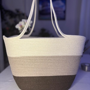Puede incluir: Un bolso tote confeccionado con cuerda trenzada que presenta un diseño de bloques de color con tres secciones horizontales. La parte superior es blanco roto, la central es beige claro y la base es marrón oscuro. Incluye dos asas largas de cuerda.