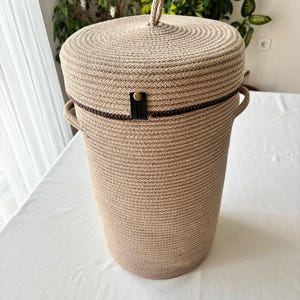 May include: A tall, cylindrical storage basket crafted from coiled beige rope. It features a matching lid with a small rope handle at the top, two side handles, and a dark brown leather-like tab secured with a gold-toned metal rivet on the lid.