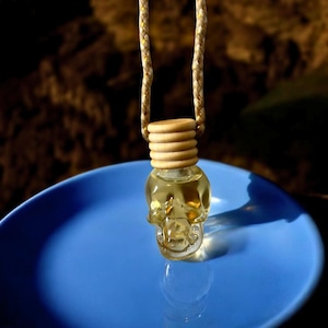 Puede incluir: Una botella de vidrio transparente con forma de calavera llena de un líquido amarillo claro. La botella cuelga de una cuerda marrón y blanca y descansa sobre un plato azul.