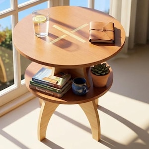 May include: A two-tiered, round wooden side table with a light brown finish. The top tier holds a glass of water with a lemon slice, and a small leather-bound notebook. The lower tier has books, a small potted plant, and a blue mug. The table is 24 inches tall.