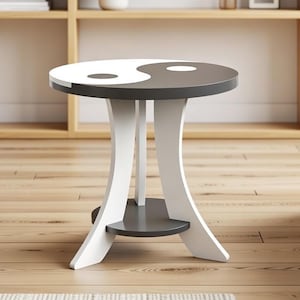 Puede incluir: Una mesa auxiliar redonda con un diseño de yin-yang en la parte superior. La parte superior está dividida en secciones blancas y gris oscuro. La mesa tiene tres patas blancas curvas y un pequeño estante gris. La mesa está en un salón.