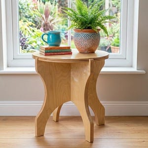 Op de afbeelding: Een kleine, lichtgekleurde houten bijzettafel met een rond blad. Een pot met varen, een blauwe mok en een stapel boeken staan op de tafel. De tafel staat bij een raam met uitzicht op een tuin.
