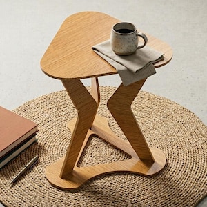 May include: A wooden side table with a unique, geometric design. The tabletop is triangular, with a mug of coffee and a folded cloth. The table is 24 inches tall and sits on a woven, circular rug. A stack of books and a pen are nearby.