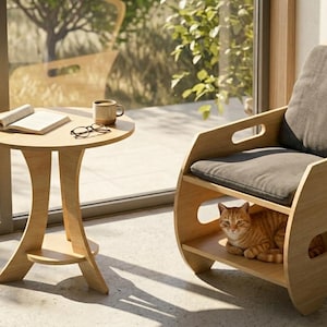 Könnte beinhalten: Ein heller Holzstuhl mit grauem Kissen und passendem Beistelltisch. Eine orangefarbene getigerte Katze sitzt im unteren Regal des Stuhls. Ein Buch, eine Brille und eine Tasse stehen auf dem Tisch.