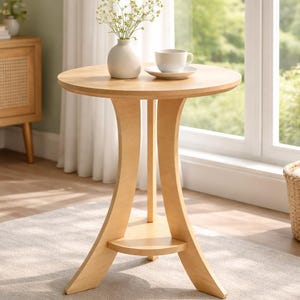 Puede incluir: Una mesa auxiliar redonda de madera clara con un pequeño estante. Un jarrón blanco con flores y una taza y platillo de té encima. La mesa tiene aproximadamente 61 cm de diámetro y 71 cm de alto.