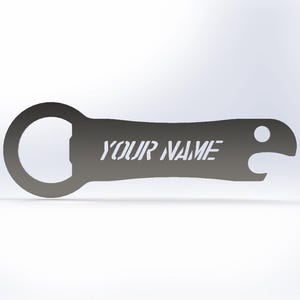 Könnte beinhalten: Ein grauer Metallflaschenöffner mit dem Text "YOUR NAME" in einer Schablonenschrift. Der Öffner hat eine kreisförmige Öffnung für den Flaschenverschluss und einen kleinen Haken zum Entfernen des Verschlusses. Das Design ist schlicht und modern.