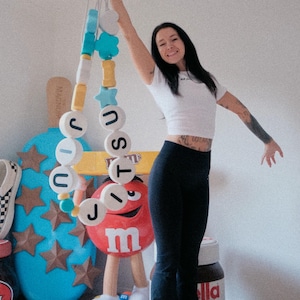 May include: A long decorative garland featuring large, round white beads with black letters spelling out JIU JITSU, interspersed with smaller, colourful star-shaped and cylindrical beads in shades of blue, yellow, and white, suspended on a white cord.