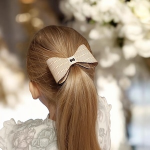 May include: A light brown wooden bow hair clip with a vertical striped pattern and a small black center piece. The bow is attached to a ponytail of long, light brown hair. The background is blurred with white flowers.