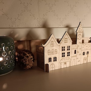May include: A wooden decorative cityscape featuring laser-cut buildings with windows, doors, and a clock tower. A dark green ceramic candle holder with circular cutouts and a lit candle sits to the left, along with pine cones.