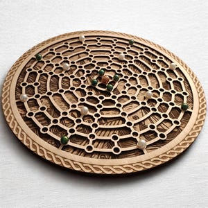 Op de afbeelding: Een houten bordspel met een cirkelvormig ontwerp. Het bord heeft een netwerk van onderling verbonden gaten en een decoratieve rand. Groene, witte en bruine knikkers zijn verspreid over het bord. Het spel is gemaakt van lichtgekleurd hout.