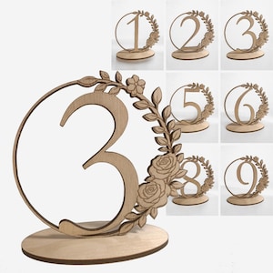 Puede incluir: Números de mesa de madera, del 1 al 9, ideales para eventos. Cada número está cortado con láser en madera clara, con un diseño de corona floral. Los números se apoyan en una base redonda, añadiendo un toque rústico a la decoración.