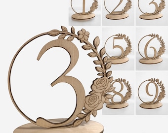 May include: Wooden table numbers 1-9, perfect for weddings or events. Each number is laser-cut from light-colored wood, featuring a floral wreath design. The numbers stand on a round base, adding a touch of rustic elegance to any table setting.