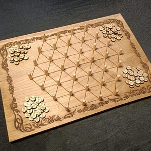Puede incluir: Un tablero de juego de madera con un patrón de cuadrícula y pequeñas clavijas de madera. También hay pequeñas fichas de color dorado esparcidas por el tablero.