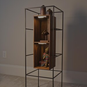 May include: A tall, dark metal display shelf with a wooden cabinet. The cabinet has two shelves with a small statue and decorative items. The shelf is freestanding and has a minimalist design, suitable for displaying collectibles or decor.