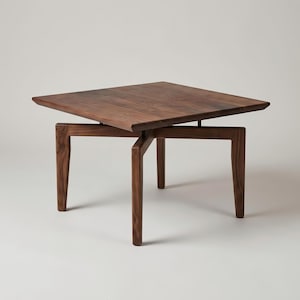 May include: A square coffee table crafted from dark brown wood with a visible grain pattern. The table features a flat top surface supported by four angled legs that intersect in a cross-like structure beneath the center of the tabletop.