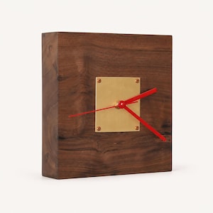 May include: A square wall clock featuring a dark brown wood grain body. A square brass plate is centered on the front, secured by four small screws. Three bright red clock hands are attached to the center of the brass plate, indicating the time.