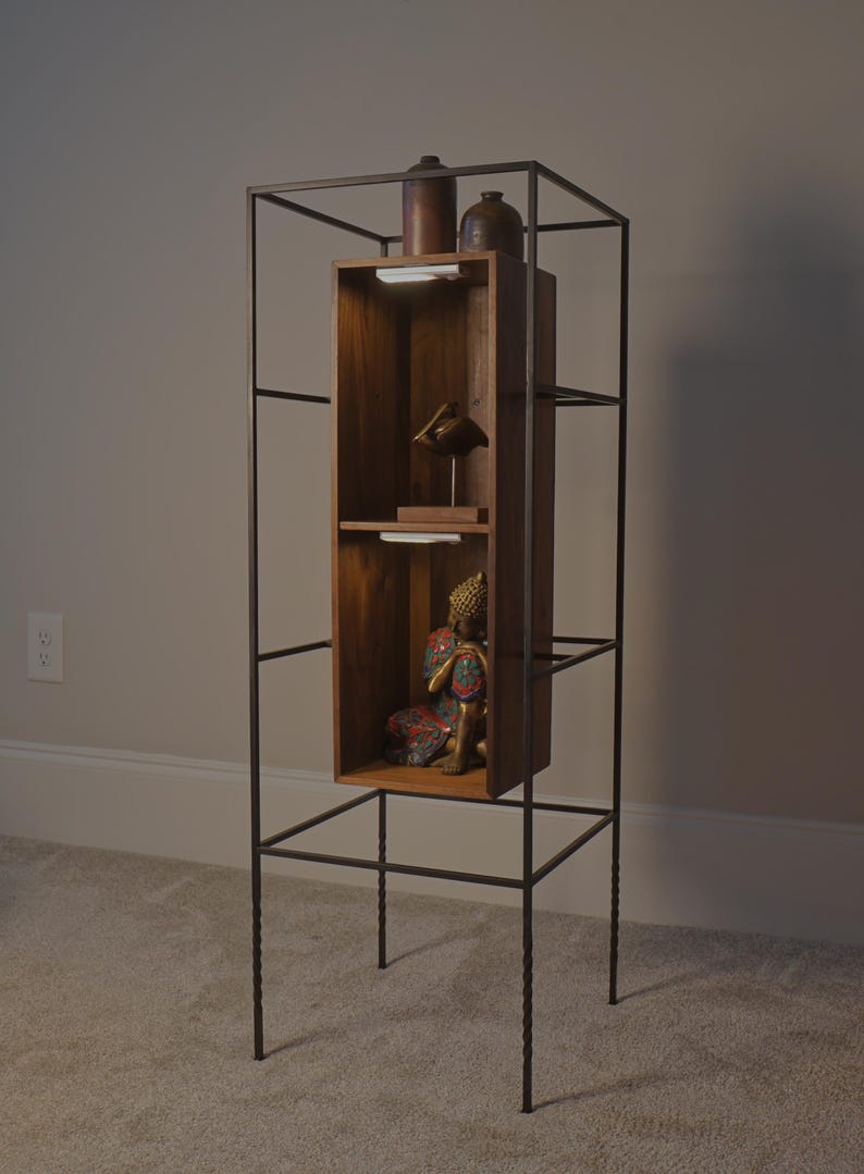 May include: A tall, dark metal display shelf with a wooden cabinet. The cabinet has two shelves with a small statue and decorative items. The shelf is freestanding and has a minimalist design, suitable for displaying collectibles or decor.