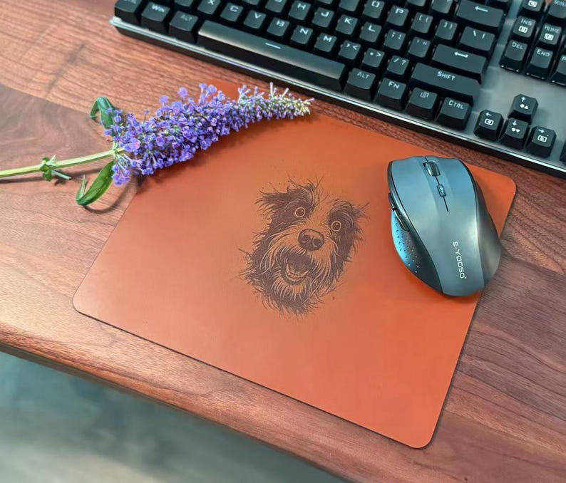 Pode incluir: Um tapete de rato laranja queimado com uma ilustra&ccedil;&atilde;o detalhada do rosto de um c&atilde;o, um rato sem fios, um teclado e um raminho de flores roxas. O tapete &eacute; retangular e est&aacute; numa secret&aacute;ria de madeira.