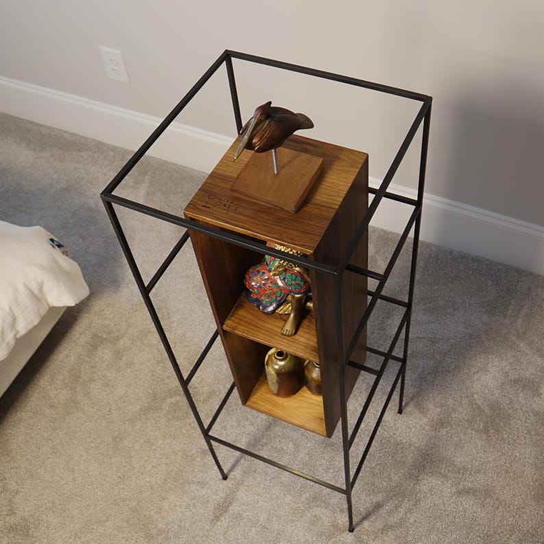 May include: A tall, black metal and wood display shelf with a pelican sculpture on top. The shelf has a rectangular wooden box with shelves inside, holding decorative items. The metal frame surrounds the wooden box.