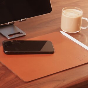 Pode incluir: Um tapete de mesa de couro castanho com um smartphone preto, uma caneta stylus branca e uma caneca de caf&eacute; de vidro. Um suporte de tablet prateado e um monitor de computador est&atilde;o no fundo. O tapete tem o texto "Leather Lines" em relevo.