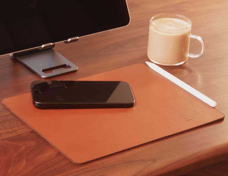 Pode incluir: Um tapete de mesa de couro castanho com um smartphone preto, uma caneta stylus branca e uma caneca de caf&eacute; de vidro. Um suporte de tablet prateado e um monitor de computador est&atilde;o no fundo. O tapete tem o texto "Leather Lines" em relevo.
