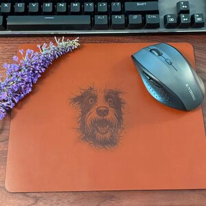 Pode incluir: Um mousepad laranja queimado com uma ilustra&ccedil;&atilde;o detalhada do rosto de um c&atilde;o. Um mouse sem fio e um raminho de flores roxas tamb&eacute;m est&atilde;o no mousepad. Um teclado preto est&aacute; ao fundo.