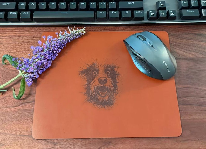 Pode incluir: Um mousepad laranja queimado com uma ilustra&ccedil;&atilde;o detalhada do rosto de um c&atilde;o. Um mouse sem fio e um raminho de flores roxas tamb&eacute;m est&atilde;o no mousepad. Um teclado preto est&aacute; ao fundo.