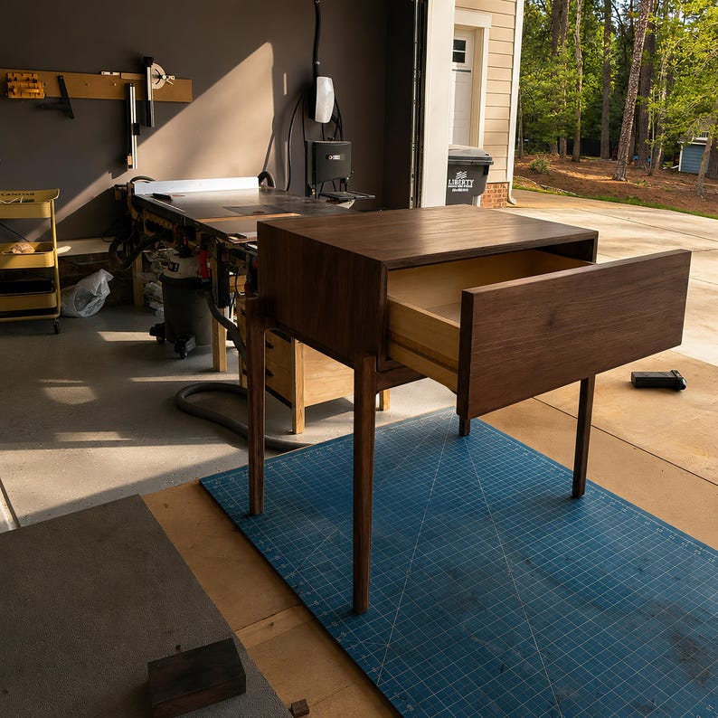 Puede incluir: Una mesa auxiliar de madera de color marr&oacute;n oscuro con cuatro patas delgadas y un &uacute;nico caj&oacute;n horizontal ancho, abierto para revelar un interior de color claro. La mesa presenta una superficie superior rectangular lisa y un marco limpio.