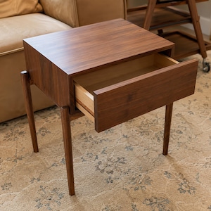 Puede incluir: Una mesa auxiliar de madera de color marr&oacute;n oscuro con una superficie rectangular y un caj&oacute;n extra&iacute;ble. La mesa cuenta con cuatro patas delgadas y c&oacute;nicas, y un patr&oacute;n de veta de madera visible. El caj&oacute;n est&aacute; entreabierto, mostrando un interior claro.